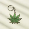 Green marijuana leaf keychain resting on a white surface, showcasing its vibrant color and detailed leaf design.