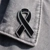 A black awareness ribbon lapel pin with a silver outline, attached to the collar of a gray denim jacket, symbolizing remembrance and support.