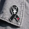 A black ribbon pin featuring a white cross at the top and a red heart near the bottom. The pin is attached to a grey fabric, combining religious and emotional symbols in its design.
