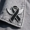 A black ribbon pin with silver wings attached, placed on a denim fabric. The pin symbolizes awareness, with a sleek and modern design.
