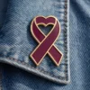 A burgundy ribbon pin shaped with a heart at the top, outlined in gold. The pin is displayed on the collar of a denim jacket, offering a soft glossy finish.