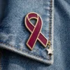 A burgundy ribbon pin with a glossy finish, outlined in gold, featuring a small silver star charm hanging from the bottom. The pin is displayed on the collar of a denim jacket.