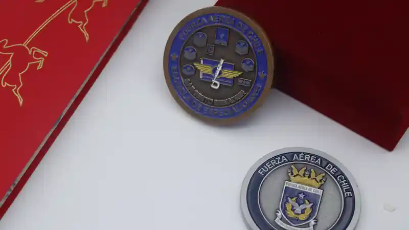 Dos monedas de desafío de la Fuerza Aérea de Chile. La moneda de bronce superior lleva inscrito "FUERZA AÉREA DE CHILE" y "ESCUELA SARGENTO MENADIER DE ESPECIALIDADES". En la moneda de plata inferior figura el emblema de la Fuerza Aérea de Chile y "FUERZA AÉREA DE CHILE"."