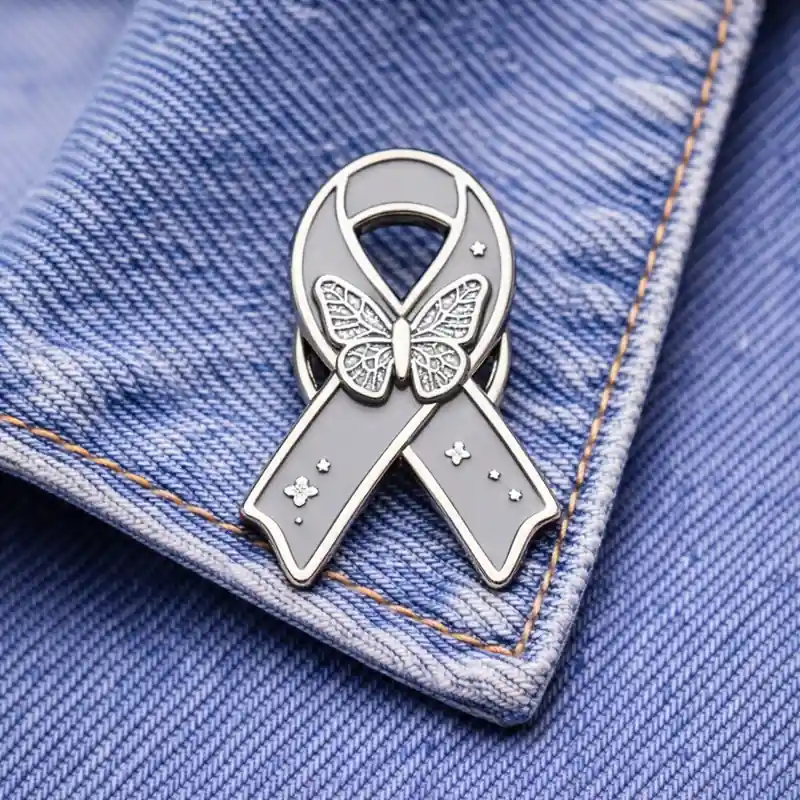 A custom gray ribbon enamel pin with a butterfly design and stars, displayed on a denim fabric background.
