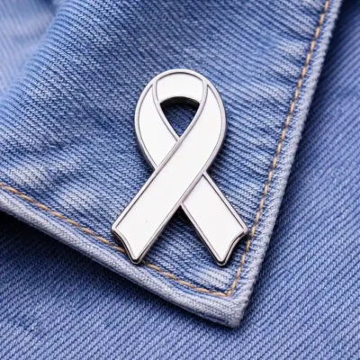A custom white ribbon enamel pin with space for personalized text at the bottom, displayed on a denim fabric.