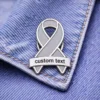 A gray ribbon enamel pin with a customizable text banner, displayed on denim fabric.