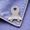 A gray enamel ribbon pin with a customizable "custom text" banner, pinned to denim fabric.