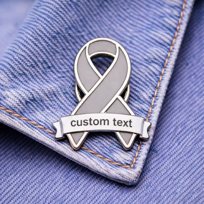 A gray enamel ribbon pin with a customizable "custom text" banner, pinned to denim fabric.