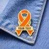 Customizable orange awareness ribbon enamel pin with personalized text, displayed on denim fabric, representing support for fire-related causes or charity events.