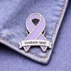 A purple enamel ribbon pin with a customizable "custom text" banner, pinned to denim fabric.