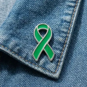 Green awareness ribbon enamel pin displayed on a blue denim jacket, symbolizing support for health awareness and charity causes.
