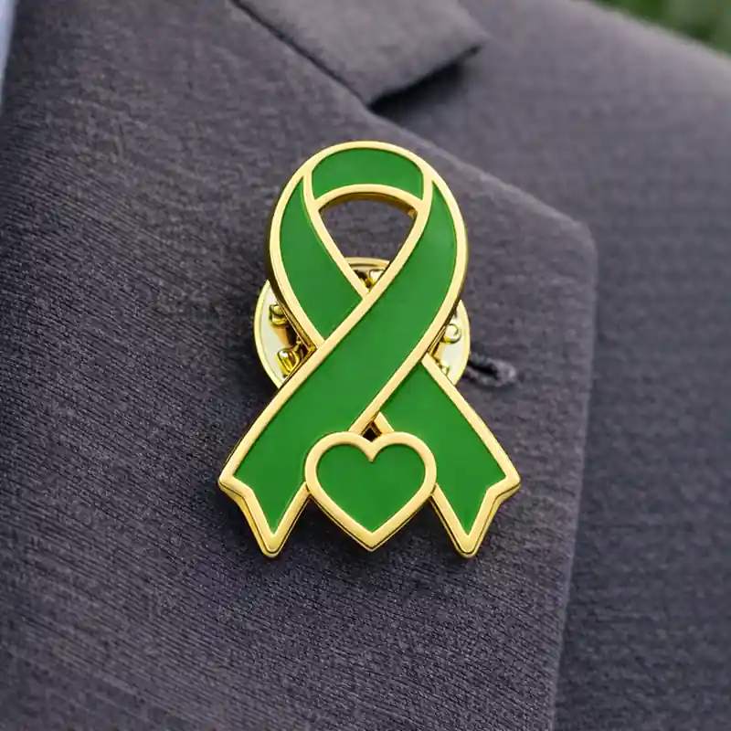 Green awareness ribbon enamel pin with gold outline and heart detail, worn on a suit jacket to symbolize support for health awareness and social causes.