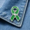 Green awareness ribbon enamel pin with silver heart detail, worn on a blue denim jacket to support health awareness and charity causes.