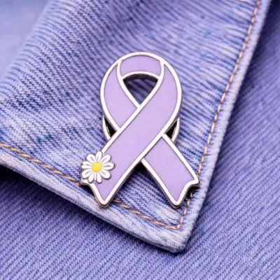A lavender enamel ribbon pin with silver edges and a white daisy design attached to a denim jacket.