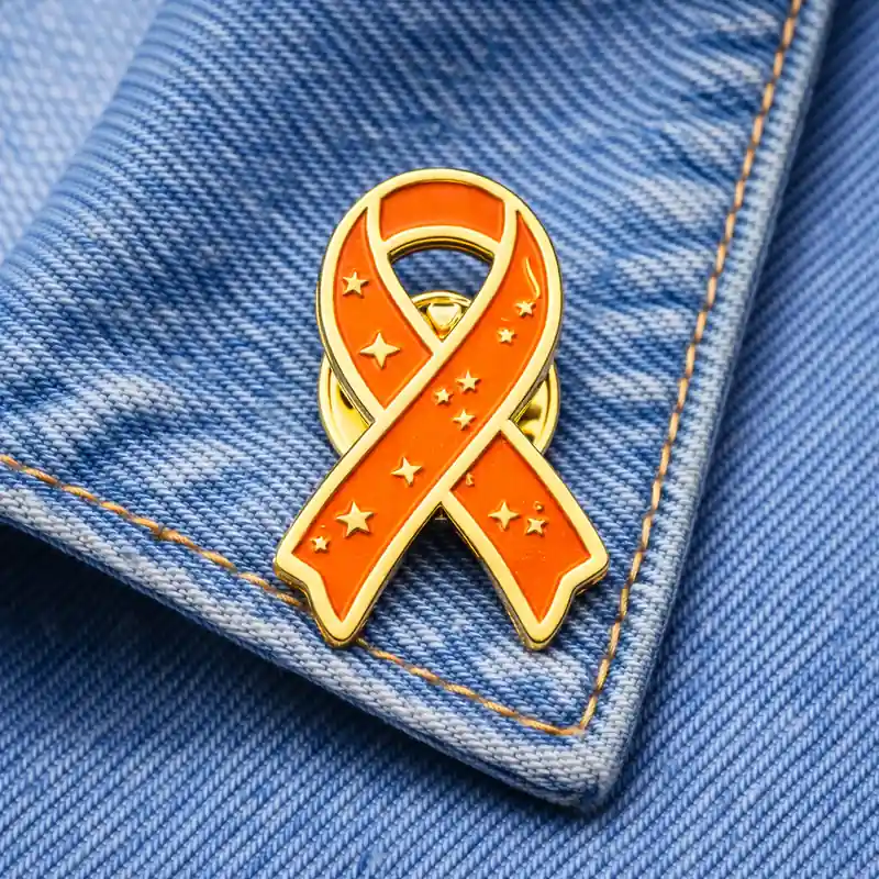 Orange awareness ribbon enamel pin with stars design, displayed on denim fabric, symbolizing support for fire-related causes or awareness campaigns.