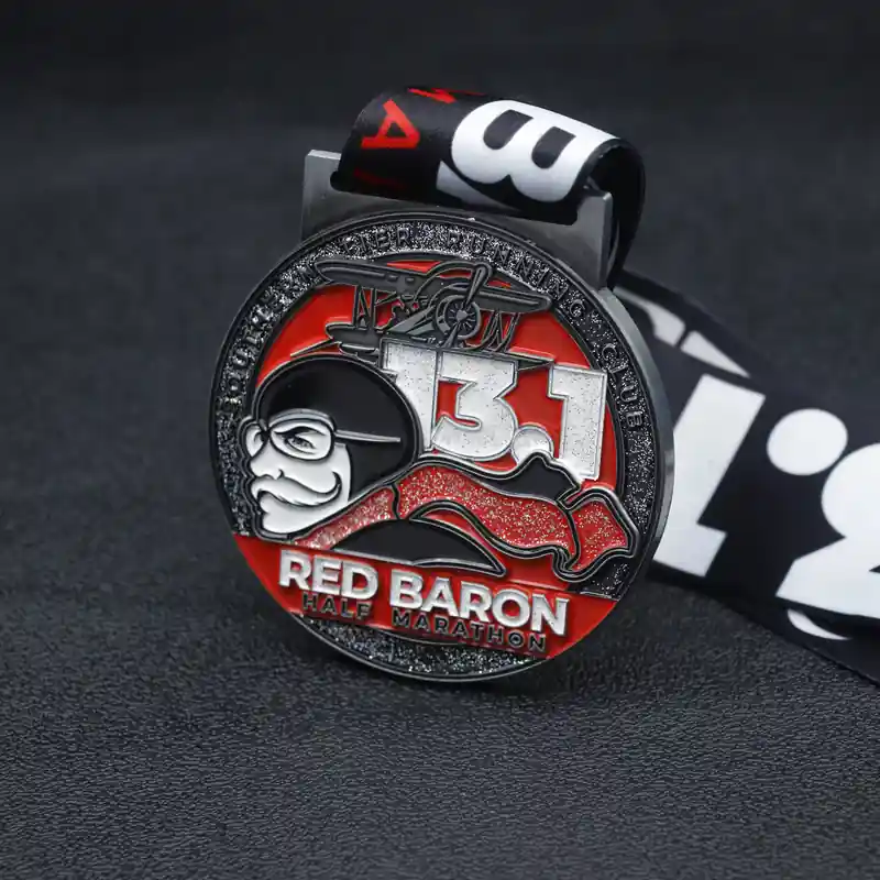 Close-up image of the Red Baron Half Marathon medal with a red and silver design featuring the number "13.1" and an airplane graphic. The medal is attached to a black and white ribbon with the event name visible at the bottom. The medal has a glittery red finish and is placed on a dark surface.
