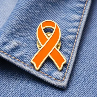 Solid orange awareness ribbon enamel pin, displayed on denim fabric, symbolizing support for fire-related causes, awareness campaigns, or charitable initiatives.