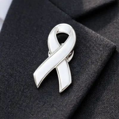 A white ribbon pin with silver edging, placed on a black fabric background. The pin's smooth, glossy surface contrasts with the dark fabric.