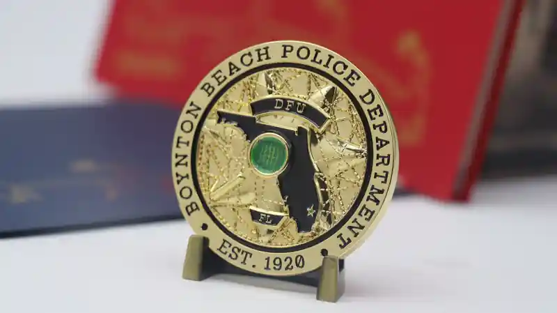 Moneda de desafío de oro del Departamento de Policía de Boynton Beach. En el canto de la moneda puede leerse "BOYNTON BEACH POLICE DEPARTMENT EST. 1920". El centro muestra el contorno de Florida, con un patrón de código binario sobre la ubicación de Boynton Beach, y las letras "DFU" y "FL".