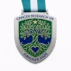 A silver and green medal for Cancer Research UK, featuring a tree design with roots and branches forming a heart shape. The tree is framed with blue and green elements, and the top has the inscription "CANCER RESEARCH UK" and "FINISHER 2026" at the bottom. The ribbon is teal and white.