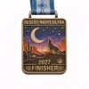 A bronze Desert Night Ultra finisher medal featuring a desert landscape under a starry sky with a crescent moon. A lone wolf howls towards the sky, and tall cacti and rocky formations surround the scene. The medal reads "DESERT NIGHT ULTRA" at the top and "2027 FINISHER" at the bottom.