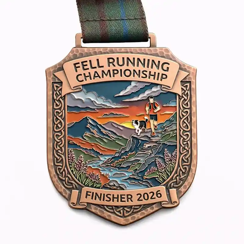 A bronze finisher medal for the Fell Running Championship 2026, showcasing a runner and a dog on a mountain trail with a vibrant sunset in the background. The medal has Celtic knotwork designs along the edges, and the top inscription reads "FELL RUNNING CHAMPIONSHIP" with "FINISHER 2026" at the bottom.