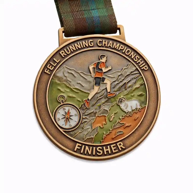 A bronze finisher medal for the Fell Running Championship featuring an image of a runner climbing a mountain, with a compass and a sheep in the foreground. The top of the medal reads "FELL RUNNING CHAMPIONSHIP," and the bottom says "FINISHER."