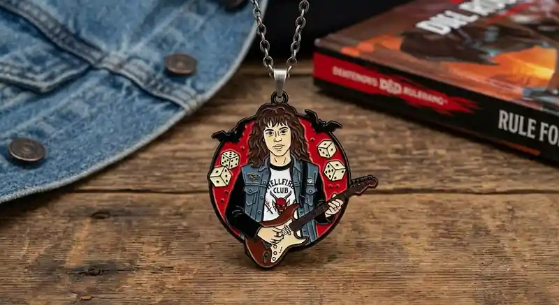 Un collar con colgante esmaltado que representa a un guitarrista de rock inspirado en la estética del metal de los 80, sosteniendo una guitarra eléctrica y vistiendo una camiseta del Hellfire Club, expuesto sobre una mesa de madera con una chaqueta vaquera y un libro al fondo.