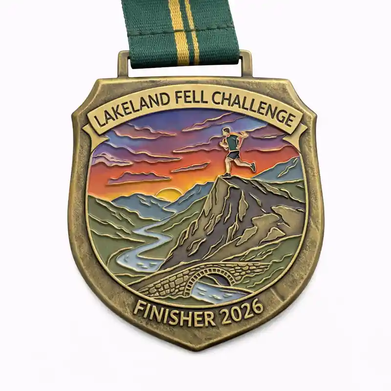 A bronze finisher medal for the Lakeland Fell Challenge 2026, showcasing a runner atop a mountain with a sunset sky in the background. The landscape includes a river, a stone bridge, and rolling hills. The medal is inscribed with "LAKELAND FELL CHALLENGE" and "FINISHER 2026," with a green and yellow ribbon.
