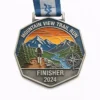 A silver Mountain View Trail Run finisher medal, showcasing a hiker running along a mountain trail with towering peaks, a river, and a setting sun in the background. The medal is inscribed with "MOUNTAIN VIEW TRAIL RUN" at the top, "FINISHER" and "2024" at the bottom, with a compass marking the directions.