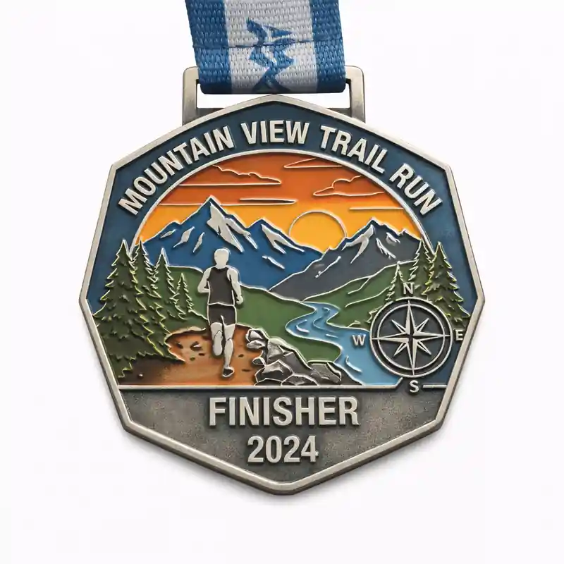A silver Mountain View Trail Run finisher medal, showcasing a hiker running along a mountain trail with towering peaks, a river, and a setting sun in the background. The medal is inscribed with "MOUNTAIN VIEW TRAIL RUN" at the top, "FINISHER" and "2024" at the bottom, with a compass marking the directions.