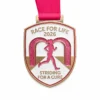A shield-shaped medal with a pink runner silhouette in front of glittery pink arches. The words "RACE FOR LIFE 2026" appear at the top, and "STRIDING FOR A CURE" is written at the bottom. The medal is attached to a pink ribbon.