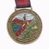 A bronze medal for the Scottish Highlands Trail Run, featuring a runner in a kilt traversing a mountainous landscape with a lake and castle in the background. The medal has Celtic knotwork around the border, and the inscription "SCOTTISH HIGHLANDS TRAIL RUN" and "FINISHER 2018" at the bottom. The ribbon is red and green plaid.