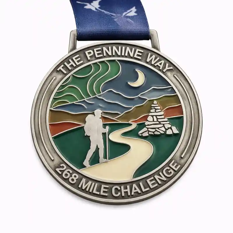 A silver and enamel medal for The Pennine Way 268 Mile Challenge, featuring an image of a hiker walking along a path with a cairn and a moon in the background. The landscape shows mountains, valleys, and clouds with flowing green and orange sections. The medal has "THE PENNINE WAY" at the top and "268 MILE CHALLENGE" at the bottom. The ribbon is blue with a white mountain design.