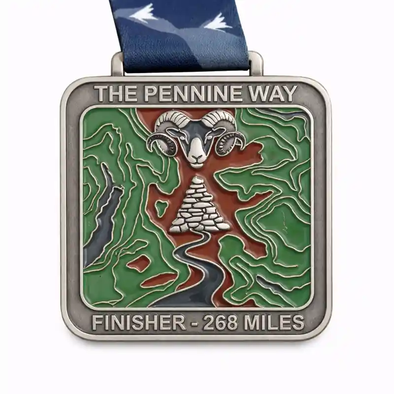 A silver medal for the Pennine Way 268 Miles featuring a design with a ram's head above a stone cairn, surrounded by a map-like pattern of green and brown terrain. A winding river flows from the cairn through the landscape. The medal is inscribed with "THE PENNINE WAY" and "FINISHER - 268 MILES," with a dark blue ribbon featuring a bird design.