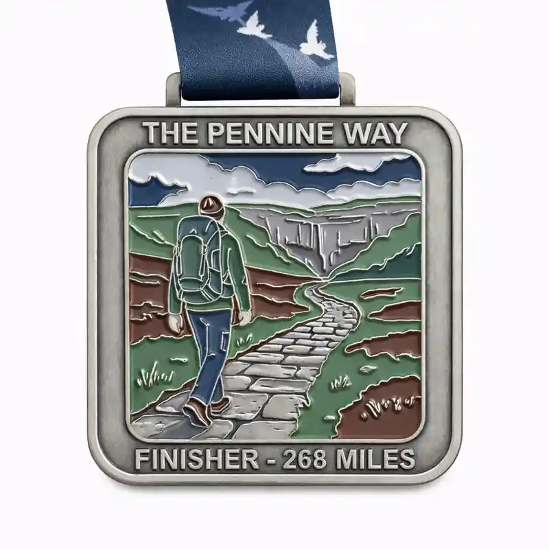 A silver medal for the Pennine Way 268 Miles featuring an image of a hiker walking along a stone path through a valley with cliffs in the distance. The scene includes rolling hills and dramatic skies. The medal is inscribed with "THE PENNINE WAY" and "FINISHER - 268 MILES," with a dark blue ribbon featuring a bird design.