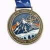 A bronze and enamel medal for "The Spine Race," a 268-mile endurance race. The medal features a hiker running across a snowy mountain range with a compass in the background. The top of the medal reads "THE SPINE RACE," and the bottom states "FINISHER - 268 MILEE." The ribbon is blue with a white bird design.