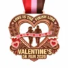 A heart-shaped rose gold medal featuring two runners crossing a finish line with the words "LOVE IS THE FINISH LINE," surrounded by chocolates and candy hearts, with the inscription "VALENTINE'S 5K RUN 2026" and a colorful ribbon attached.