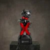 Figurine de mascotte ours noir portant des lunettes de soleil et un nœud papillon rouge, tenant une lettre X rouge en gras, montée sur un socle noir avec texte de marque