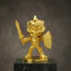 Figurine de mascotte de guerrier en or tenant une épée et un bouclier, conçue dans un style cartoon et présentée sur une base ronde au-dessus d'une plate-forme en marbre.