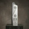 Trophée d'entreprise en argent au design géométrique angulaire moderne, gravé des logos, du nom du lauréat et d'un texte, posé sur un socle en marbre foncé