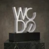 Trophée sculptural en forme de lettre WC DO en argent avec une finition moderne en métal brossé, monté sur un socle noir et présenté sur une plateforme en marbre.