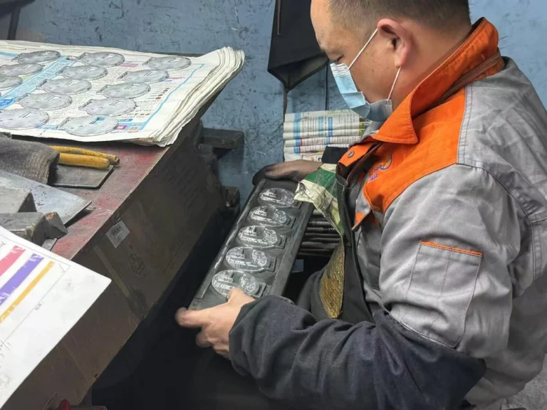 マスクを着けた工場作業員が、作業台で金属製のメダルの列を慎重に検査し、表面の品質と仕上がりを確認しながら丁寧に磨いています。