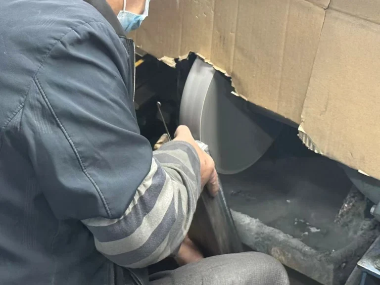 作業場で回転する研削ホイールを使って金属部品を研磨している作業員、機械の周りにはほこりや破片が見える。
