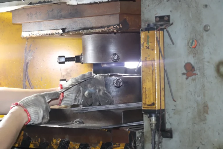 A worker wearing protective gloves operating a metal stamping press, pressing metal sheets with precise force during the manufacturing process in a factory setting.