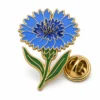 Spilla a forma di fiore di fiordaliso blu con contorni in rilievo in metallo dorato, petali blu a strati, centro scuro, gambo e foglie verdi e una farfalla in frizione su sfondo bianco.