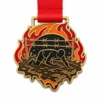 Мягкая эмалевая медаль Fire crawl Tough Mudder Medal с контурами из полированного золотистого металла, красной лентой, силуэтом ползущего бегуна, огненными дугами, грязевой лужей, соединенной линией препятствий и белым фоном.