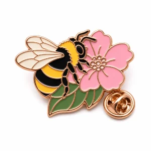 Spilla a forma di ape con contorni in metallo color oro rosa, ape gialla a strisce, ali color crema, base di fiori rosa, foglia verde e frizione a farfalla su sfondo bianco.