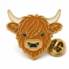 Fluffy face Highland cow pin with raised gold metal outlines, shaggy brown hair, cream horns, tan muzzle, and a butterfly clutch on white.