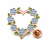 Epingle "forget me not" en forme de couronne de coeur avec des contours en métal doré rose, des fleurs émaillées bleues, des feuilles connectées vertes, un cadre en forme de coeur rose et une pochette en forme de papillon sur fond blanc.